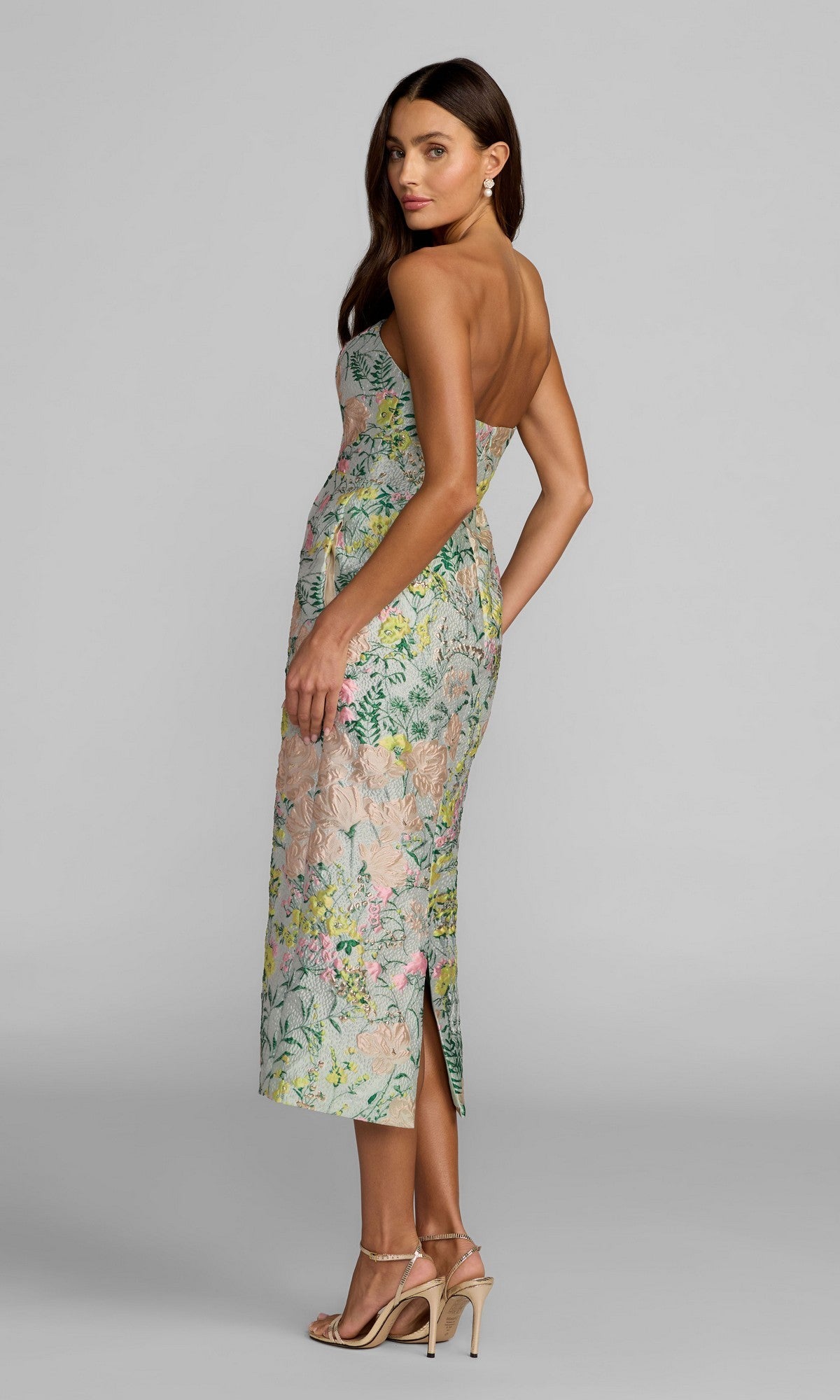 A woman wearing the Mac Duggal 13223 tea-length party dress, featuring a strapless design and delicate floral brocade pattern, stands in profile against a plain gray background, styled with strappy high heels and earrings.